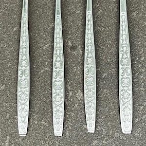 May include: Close-up of four silver forks with ornate, symmetrical patterns on the handles. The forks are arranged vertically against a textured, gray background. The tines of the forks are visible at the top.