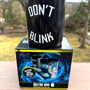 May include: Black ceramic mug with the words "DON'T BLINK" in white. The mug sits on a box with a space-themed design and the words "DOCTOR WHO" and "WEEPING ANGELS HEAT REVEAL MUG". The box also features the text "WEEPING ANGEL APPEARS WHEN HOT WATER IS ADDED".