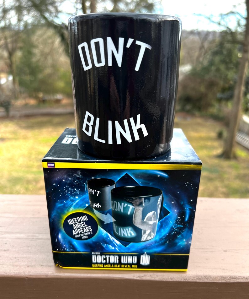 May include: Black ceramic mug with the words "DON'T BLINK" in white. The mug sits on a box with a space-themed design and the words "DOCTOR WHO" and "WEEPING ANGELS HEAT REVEAL MUG". The box also features the text "WEEPING ANGEL APPEARS WHEN HOT WATER IS ADDED".