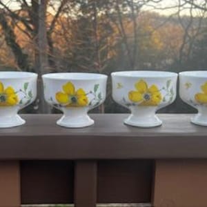 May include: Six white footed dessert bowls. Each bowl features a yellow flower design with green leaves. The bowls are arranged in a row on a brown surface, with a blurred background of trees and sky.