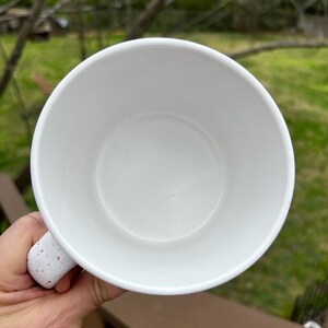 May include: White ceramic mug with a speckled handle. The interior of the mug is white and empty. The handle has red speckles. The mug is held by a hand.