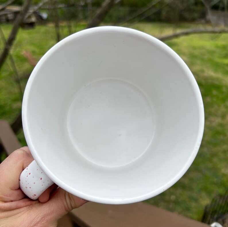 May include: White ceramic mug with a speckled handle. The interior of the mug is white and empty. The handle has red speckles. The mug is held by a hand.