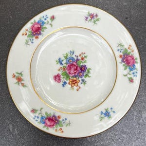 May include: A white porcelain plate with a gold rim and floral design. The plate features a central bouquet of pink, blue, and yellow flowers, with additional floral clusters around the edge. The flowers are detailed and vibrant.