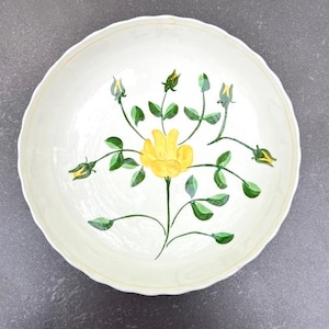 May include: A white ceramic bowl with a scalloped rim features a hand-painted floral design. The design includes a yellow rose with green leaves and stems.