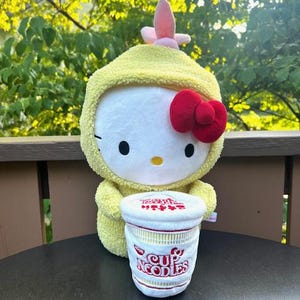 May include: A plush Hello Kitty doll wearing a yellow hooded outfit with a pink flower and red bow. The doll is holding a plush Cup Noodles container with red and white text.