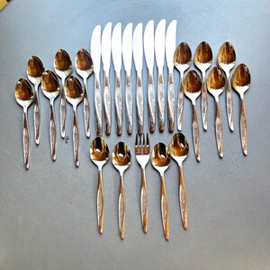 May include: A collection of vintage silverware, including spoons, knives, and forks, arranged on a gray surface. The spoons and forks have a gold finish, while the knives have silver blades. The handles feature a decorative etched design.