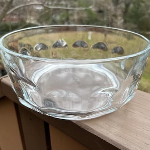 May include: A clear glass bowl with a textured, wavy design. The rim features a series of small, rounded indentations. The bowl is sitting on a wooden surface, with a blurred outdoor background.