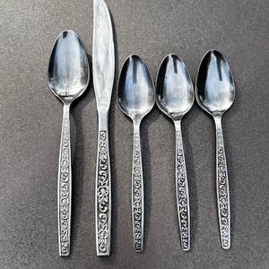 May include: A set of five silver-colored utensils: four spoons and one knife. The handles feature an ornate floral design. The utensils are arranged on a dark gray surface, reflecting the light.