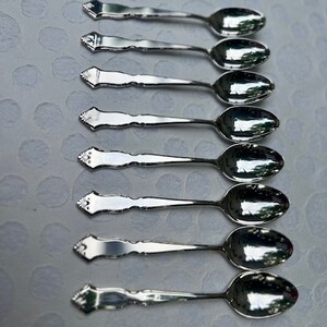 May include: Eight silver teaspoons with ornate handles. The spoons are arranged in a row, with the handles pointing to the right. The spoons are all the same size and shape.