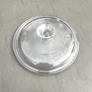 May include: A clear glass lid with a circular design. The lid features a central knob and a series of concentric ridges radiating outwards. The lid is designed to fit on a pot or pan.