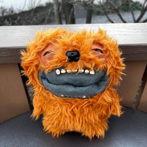 May include: A plush orange monster with a shaggy fur texture. It has a gray mouth with white teeth, a black nose, and orange eyes. The handmade creature is sitting on a dark surface.