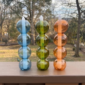 May include: Three decorative glass candle holders in blue, green, and orange. Each holder is composed of four stacked, spherical glass bulbs. The blue holder has a vertical blue stripe, the green has a green stripe, and the orange has an orange stripe.