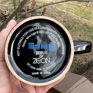 May include: Black ceramic mug with a handle, featuring the Doctor Who logo and text. The bottom of the mug has the text "NOT SUITABLE FOR USE IN A DISHWASHER OR MICROWAVE". The mug is marked "MADE IN CHINA".