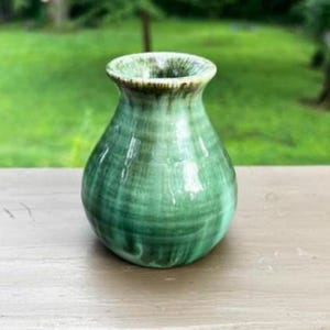 May include: A small, green ceramic vase with a rounded body and a flared rim. The vase has a glossy finish with darker green streaks, creating a marbled effect. The rim is a darker shade of green. The vase is sitting on a wooden surface.
