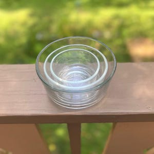 May include: A set of three clear glass bowls nested inside each other. The bowls are round with a simple design. The bowls are sitting on a wooden surface with a blurred green background.