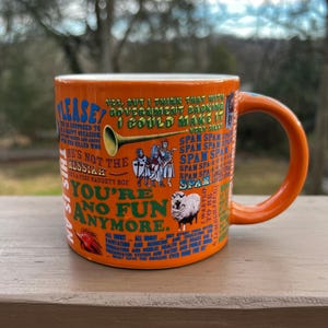 May include: Orange ceramic mug with a matching handle. The mug features various text and illustrations, including a sheep, a knight, and the word "SPAM" repeated multiple times. The mug has a white interior.