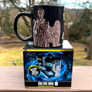 May include: Black ceramic mug featuring a Weeping Angel design from Doctor Who. The mug has a black handle and is displayed on top of its original box. The box features the Doctor Who logo and the words "Weeping Angels Heat Reveal Mug."