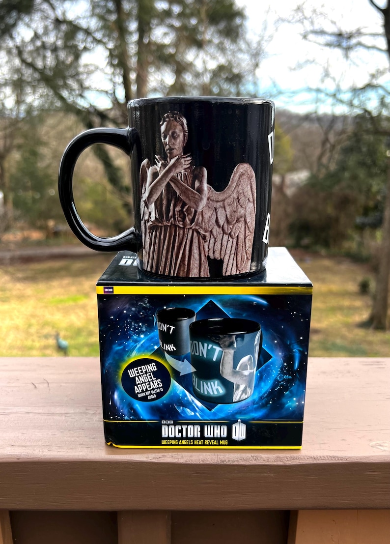 May include: Black ceramic mug featuring a Weeping Angel design from Doctor Who. The mug has a black handle and is displayed on top of its original box. The box features the Doctor Who logo and the words "Weeping Angels Heat Reveal Mug."