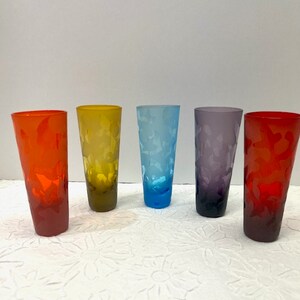May include: Five tall, colorful drinking glasses. The glasses are in shades of red, yellow, blue, purple, and red. Each glass has a textured surface. The glasses are arranged in a row against a white background.