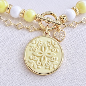 May include: A gold-toned toggle clasp necklace featuring alternating pale yellow and white spherical beads with gold spacers. A circular pendant with a raised floral motif hangs from the clasp, accompanied by a small heart-shaped charm with pavé stones.
