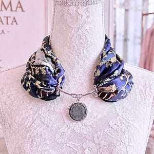 May include: A blue and gold patterned silk scarf secured by a silver-toned metal collar necklace. The necklace features a circular silver pendant with an embossed architectural design hanging from the center of the scarf and metal hardware.