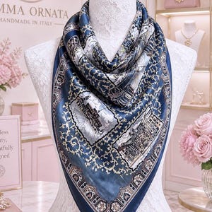 May include: A square silk scarf featuring a deep blue background with intricate gold filigree patterns and detailed architectural illustrations. The scarf is draped around a white lace-covered mannequin, showcasing a symmetrical design with borders.
