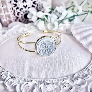 May include: Gold-tone cuff bracelet featuring a circular, light blue cameo with a relief of the Colosseum and the word "ROME". The bracelet is displayed on a white, decorative surface with floral accents.