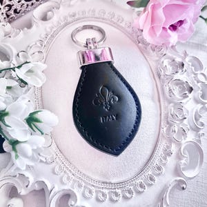 Op de afbeelding: Zwarte leren sleutelhanger met een zilverkleurige metalen ring en accenten. De sleutelhanger heeft een reliëf fleur-de-lis ontwerp en het woord "ITALY". De sleutelhanger is geplaatst tegen een witte sierlijke lijst met roze bloemen.