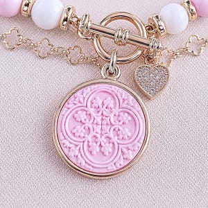 Könnte beinhalten: Ein Armband mit goldenem Knebelverschluss, weißen und hellrosa Perlen sowie einer goldenen Kette mit kleeblattförmigen Gliedern. Daran befestigt sind ein runder rosa Anhänger mit geprägtem Blumenmuster und ein kleines herzförmiges Schmuckstück.