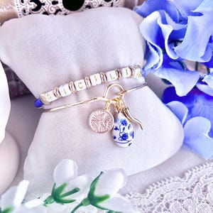 Könnte beinhalten: Ein goldfarbenes Armband mit dem Wort "SICILIA" in weißen und goldenen Perlen. Das Armband hat Charms, darunter ein Kompass, ein blau-weißes Blumen-Tröpfchen und ein goldenes Horn. Das Armband wird auf einem weißen Kissen präsentiert.
