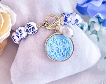 Pode incluir: Pulseira de contas azuis e brancas com fecho dourado e um pingente azul claro. O pingente tem um design floral e um pingente de pérola branca. A pulseira está sobre uma almofada branca, com flores azuis ao fundo.