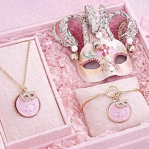 Könnte beinhalten: Eine rosa Schmuckschatulle enthält eine goldene Halskette und ein Armband, jeweils mit einem rosa Anhänger und einem Maskenmuster. Eine dekorative Maske und eine kleine Geschenkbox sind ebenfalls enthalten. Das Set wird auf rosa Stoff präsentiert.