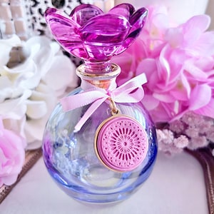 May include: A decorative glass perfume bottle with a flower-shaped stopper in shades of purple. The round bottle has a blue and purple hue, with a pink medallion charm and a pink ribbon tied around the neck. The bottle is surrounded by pink and white flowers.
