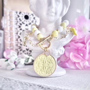 Puede incluir: Un collar dorado con un colgante redondo con un diseño floral. El collar tiene cuentas amarillas y blancas, un cierre de palanca y un pequeño dije de corazón. También hay un dije amarillo y un pequeño dije de letra.
