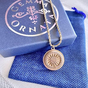 Könnte beinhalten: Goldfarbene Halskette mit einem kreisförmigen Anhänger mit Sonnenstrahlendesign und griechischem Schlüsselrand. Die Halskette liegt auf einer blauen Schachtel mit dem Markennamen "GEMMA ORNAMENTA" und einem blauen Kordelzugbeutel.