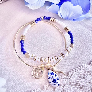 Könnte beinhalten: Goldfarbene Armreifen mit einem Armband mit weißen und blauen Perlen. Ein Armband hat den Namen "SUCHACHA" in weißen Buchstabenperlen. Zu den Anhängern gehören eine Münze und ein blau-weißes Blumen-Tröpfchen.