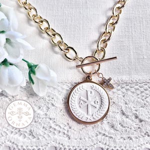 Op de afbeelding: Goudkleurige ketting met een witte hanger met een religieus symbool en een kleine kruisbedel. De hanger is rond met een verhoogd ontwerp en een goudkleurige rand. De ketting heeft een tuimelsluiting.