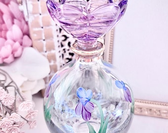 May include: A clear, spherical glass perfume bottle featuring hand-painted purple and blue iris flowers with green stems. The bottle is topped with a decorative, translucent purple glass stopper shaped like a flower blossom and a gold-toned neck.