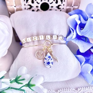 May include: Two bracelets displayed on a white cushion. One bracelet spells "SICILIAN" with white letter beads. The other is gold with a coin charm, a blue and white teardrop charm, and a gold horn charm. Blue flowers are in the background.