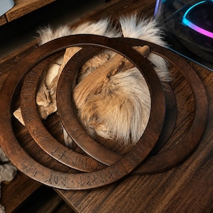 May include: Three dark brown circular wooden rings featuring engraved runic characters along their surfaces. The rings are layered over a piece of light tan, textured animal fur, creating a contrast between the smooth wood and the soft material.