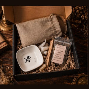 May include: A black box with a kraft paper interior holds a small white ceramic bowl with black symbols, a burlap bag, a small bag with text, a bundle of sticks, and small crystals. The text on the bag reads "Set the offering".