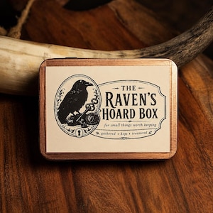 May include: A rectangular copper-colored metal tin featuring a cream-colored label with a black illustration of a raven perched on a key. The text reads: THE RAVEN'S HOARD BOX, for small things worth keeping, gathered, kept, treasured.