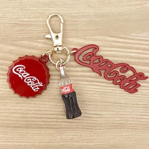 May include: Red and gold Coca-Cola keychain with a bottle charm, a bottle cap charm, and a gold split ring.