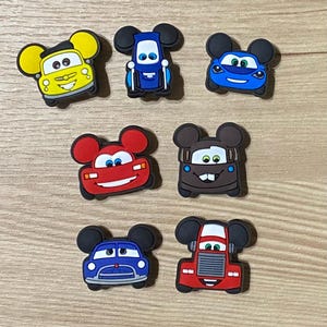 May include: Set of seven colorful cartoon car shoe charms with Mickey Mouse ears. The cars are red, blue, yellow, and brown.