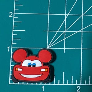 May include: Red cartoon car with Mickey Mouse ears, smiling with blue eyes. The car has headlights and a grill.