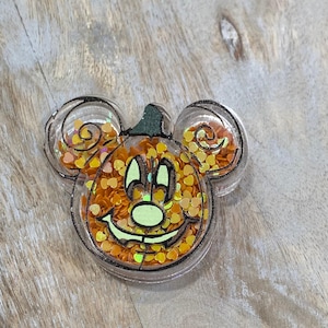 May include: A clear, Mickey Mouse-shaped shaker filled with orange and yellow heart-shaped confetti. The design features a smiling jack-o'-lantern face with a green stem. The edges are outlined in black. A festive Halloween decoration.