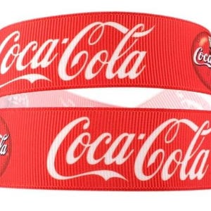May include: Red and white ribbon with the Coca-Cola logo printed on it. The ribbon is folded in half, showing the logo on both sides.