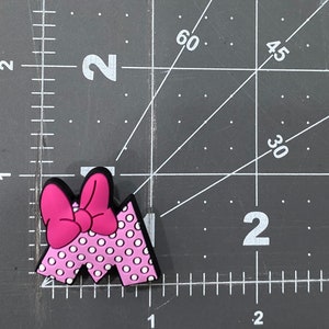 Puede incluir: Un charm con la letra "M" rosa y blanco con lunares, con un lazo rosa brillante. El charm tiene un borde negro y est&aacute; sobre una superficie negra con marcas de medici&oacute;n.