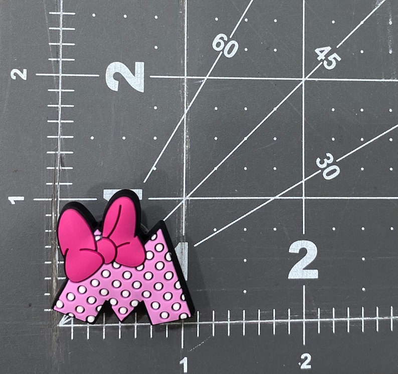 Puede incluir: Un charm con la letra "M" rosa y blanco con lunares, con un lazo rosa brillante. El charm tiene un borde negro y est&aacute; sobre una superficie negra con marcas de medici&oacute;n.