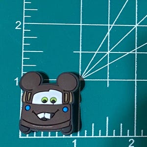 May include: Brown cartoon car with Mickey Mouse ears, green eyes, and blue accents. The car is smiling with a toothy grin.